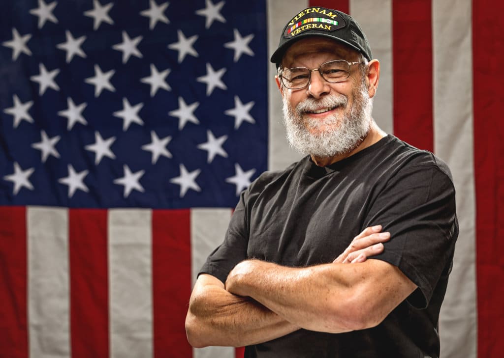 Bearded Vietnam veteran standing in front of an American Flag with arms crossed
